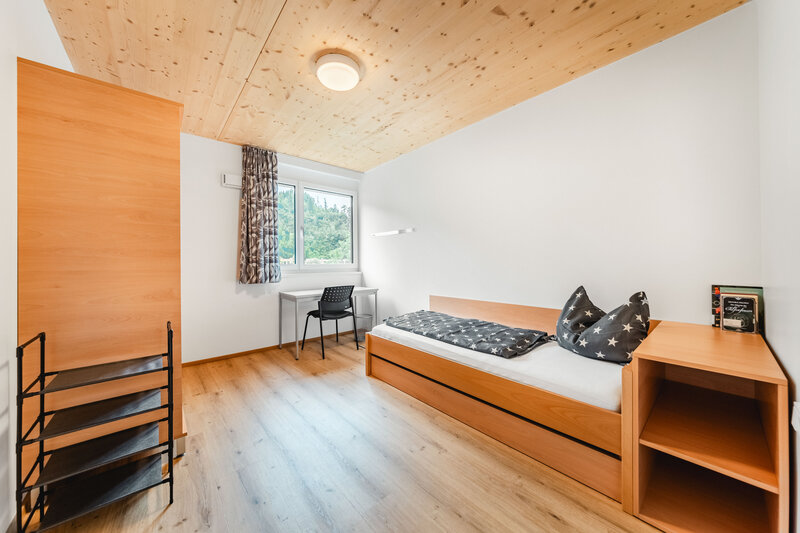 Ein helles, modern eingerichtetes Schlafzimmer mit einem Einzelbett, einer grauen Bettdecke, einem Schreibtisch mit Stuhl und einem Fenster mit Blick auf grüne Bäume. | © FH Kufstein Tirol