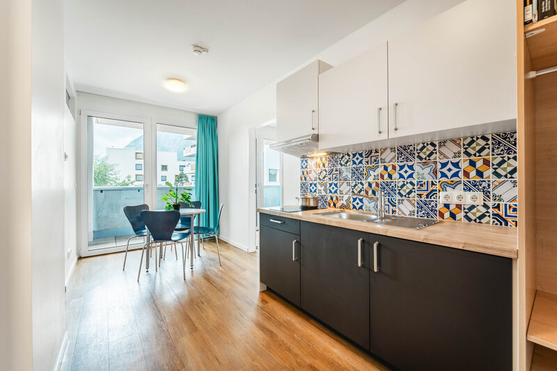 Ein helles, modernes Kücheninterieur mit glatten, dunklen Unterschränken und einer hellen Arbeitsplatte. Im Hintergrund sind ein Tisch, Stühle und ein Balkon zu sehen. | © FH Kufstein Tirol