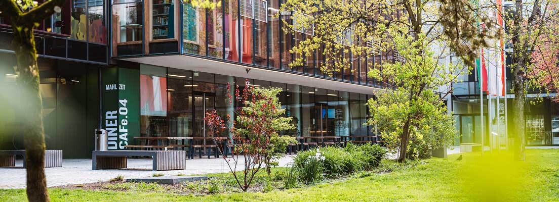 Außenansicht der FH Kufstein Tirol mit Glasfassade, Cafeteria-Bereich und Frühlingsbepflanzung auf dem Campus