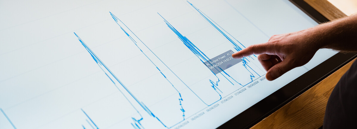 Finger zeigt auf Energiediagramm mit Leistungsdaten auf Touchscreen im Sustain & Estate Lab der FH Kufstein Tirol.