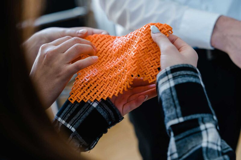 Studierende halten im Makers Lab der FH Kufstein Tirol ein flexibles, orangefarbenes 3D-gedrucktes Gewebemuster in den Händen.