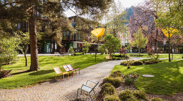 Grüner Campusbereich mit gepflastertem Weg, bunten Bänken und moderner Hochschularchitektur im Hintergrund