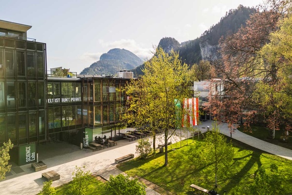 Frühlingshafte Außenaufnahme des Campus der FH Kufstein Tirol mit Glasfassade, Fahnen und Bergkulisse im Hintergrund | © FH Kufstein Tirol