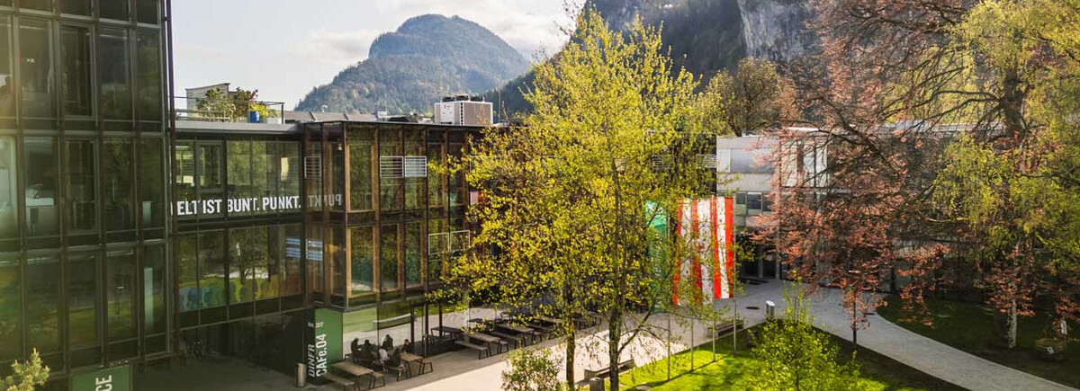 Frühlingshafte Außenaufnahme des Campus der FH Kufstein Tirol mit Glasfassade, Fahnen und Bergkulisse im Hintergrund