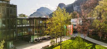 Frühlingshafte Außenaufnahme des Campus der FH Kufstein Tirol mit Glasfassade, Fahnen und Bergkulisse im Hintergrund