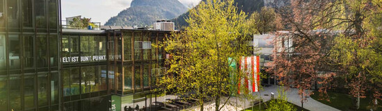 Frühlingshafte Außenaufnahme des Campus der FH Kufstein Tirol mit Glasfassade, Fahnen und Bergkulisse im Hintergrund