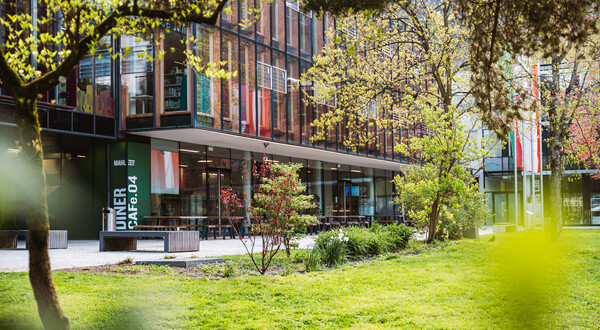 Außenansicht der FH Kufstein Tirol mit Glasfassade, Cafeteria-Bereich und Frühlingsbepflanzung auf dem Campus