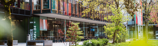 Außenansicht der FH Kufstein Tirol mit Glasfassade, Cafeteria-Bereich und Frühlingsbepflanzung auf dem Campus