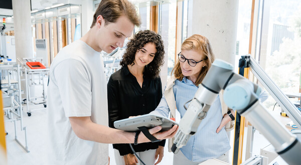 LEAN_Lab_Demonstration | © FH Kufstein Tirol Drei Personen schauen zusammen auf einen tragbaren Bildschirm, der von einem Mann gehalten wird. | © FH Kufstein Tirol