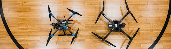 Zwei Drohnen stehen nebeneinander auf einem Hallenboden mit Spielfeldmarkierungen