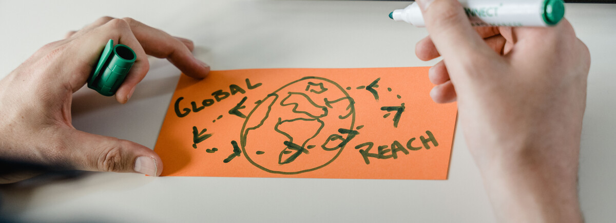 Person zeichnet eine stilisierte Weltkarte mit dem Schriftzug „Global Reach“ – Sinnbild für internationale Vernetzung, Zusammenarbeit und globale Perspektiven.