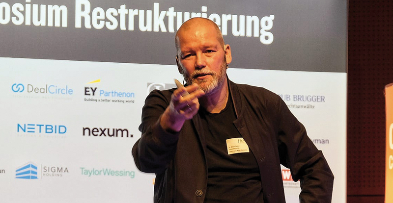Ein Mann spricht auf einer Bühne, im Hintergrund Sponsorenlogos und der Schriftzug „Symposium Restrukturierung“