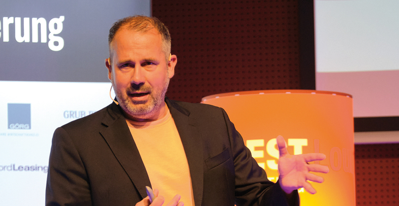 Mann steht auf einer Bühne und hält einen Vortrag, am Ohr trägt er ein Mikrophon, im Hintergrund sieht man eine Lampe und eine Tafel mit Logos