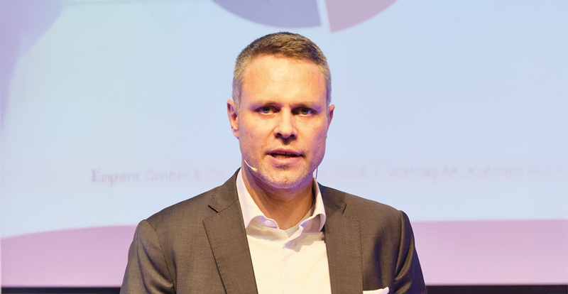 Ein Mann in Anzug und Hemd spricht auf einer Bühne und trägt ein Mikrofon am Ohr, vor einem dezenten, lilafarbenen Hintergrund