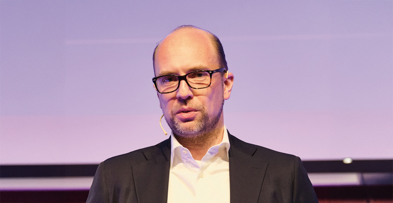 Ein Mann in Anzug und Hemd spricht auf einer Bühne und trägt ein Mikrofon am Ohr, vor einem dezenten, lilafarbenen Hintergrund