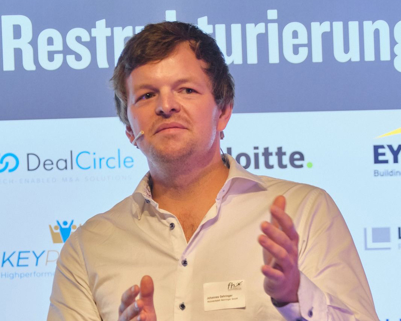 Johannes Gehringer als Speaker auf der Bühne beim 14. Symposium Restrukturierung, hinter ihn eine Wand mit der Aufschrift "Restrukturierung" und den Logos diverser Partner.