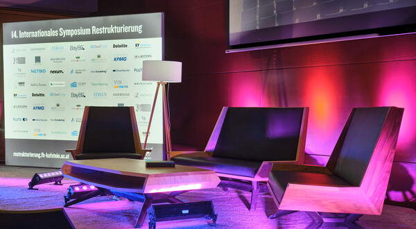 Bühne der 14. Jahreskonferenz Restrukturierung an der FH Kufstein Tirol, eine moderne Sitzgruppe und mit Tisch und Stehlampe, dahinter eine Aufsteller mit dem Schriftzug 14. Internationales Symposium Restrukturierung und vielen Logos. Die Bühne wird von lila- rosa-farbigen Scheinwerfern angestrahlt
