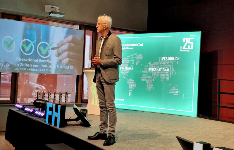Professor Martin Adam hält die Begrüßungsrede beim 24. PQM-Dialog 2026 an der FH Kufstein Tirol auf der Bühne vor einer Präsentation zu Operational Excellence.