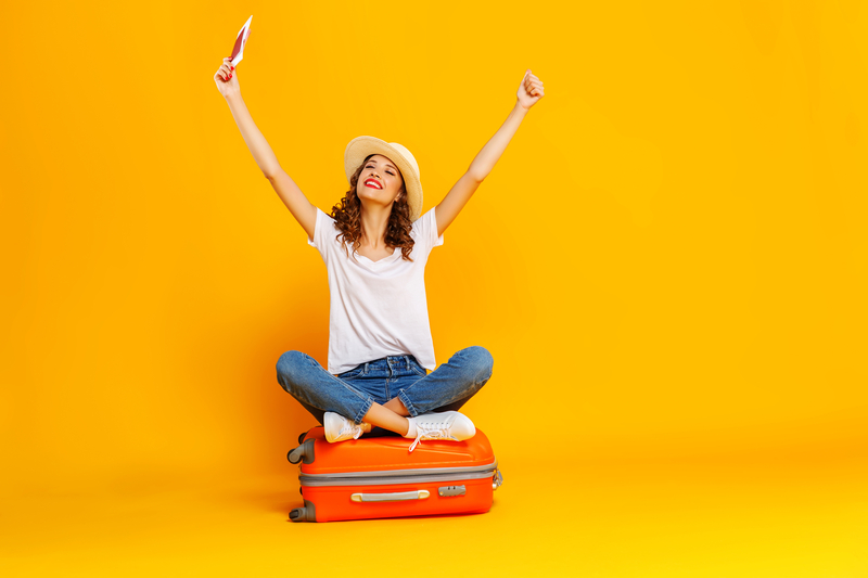 Reisebereit mit guter Laune – Mobilität und Abenteuerlust im Studium erleben Junge Frau sitzt lachend auf orangefarbenem Koffer vor farbiger Studio-Kulisse.