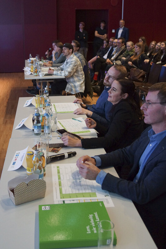 Zu sehen ist die Jury sowie das im Hintergrund befindliche Publikum der 7-Euro-Cash-Veranstaltung. Alle hören aufmerksam zu. | © FH Kufstein Tirol