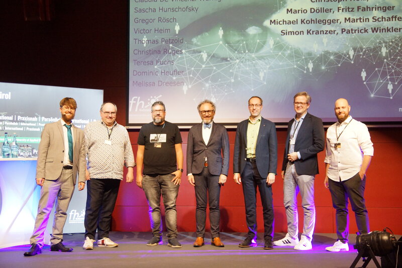 Ein Gruppenbild der Vortragenden der SPS-Tagung. Im Hintergrund befinden sich eine Werbetafel der FH Kufstein und eine Leinwand mit einer Präsentation. | © FH Kufstein Tirol