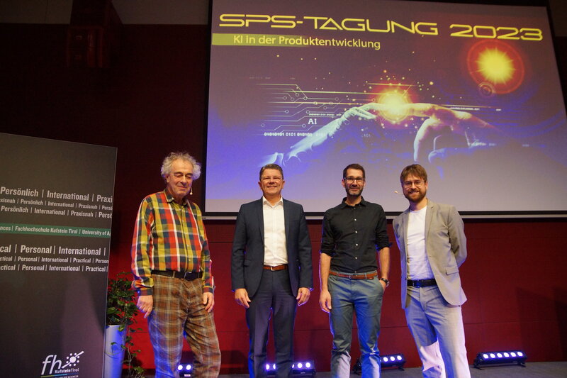 Vier Vortragende der SPS-Tagung stehen auf der Bühne und machen ein Gruppenfoto. Hinter ihnen befindet sich eine Leinwand mit einer PowerPoint-Präsentation. | © FH Kufstein Tirol