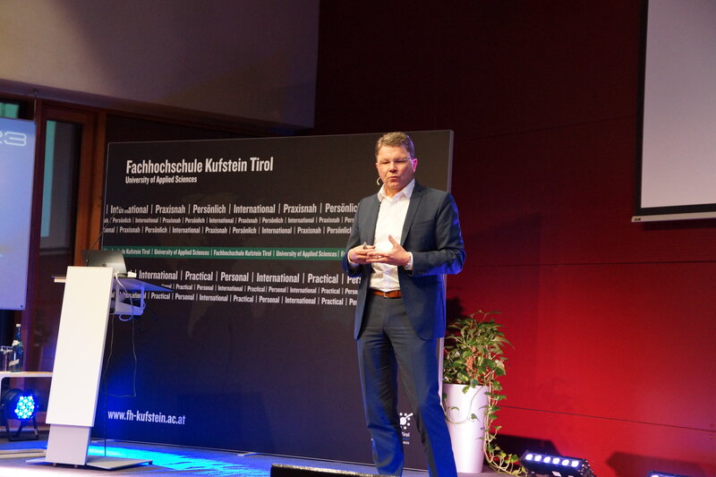 Ein Vortragender steht auf der Bühne. Hinter ihm befindet sich eine Werbetafel der FH Kufstein Tirol. | © FH Kufstein Tirol