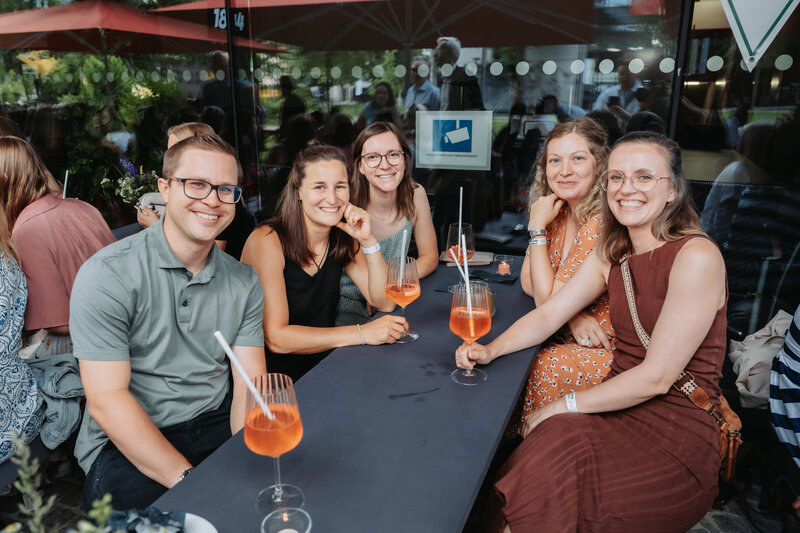 event-homecoming-gaestegruppe-tischbereich-2025 | © FH Kufstein Tirol Eine kleine Gruppe unterbricht ihr Gespräch für ein gemeinsames Lächeln in die Kamera. | © FH Kufstein Tirol