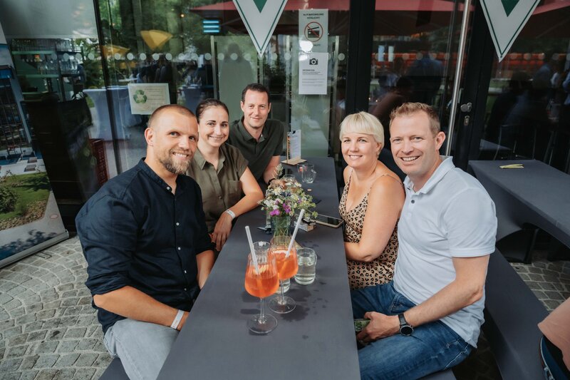 event-homecoming-gruppe-tischbereich-vor-cafe04-2025 | © FH Kufstein Tirol Eine gemischte Gruppe lacht zusammen, als sie für das Foto bemerkt wird. | © FH Kufstein Tirol
