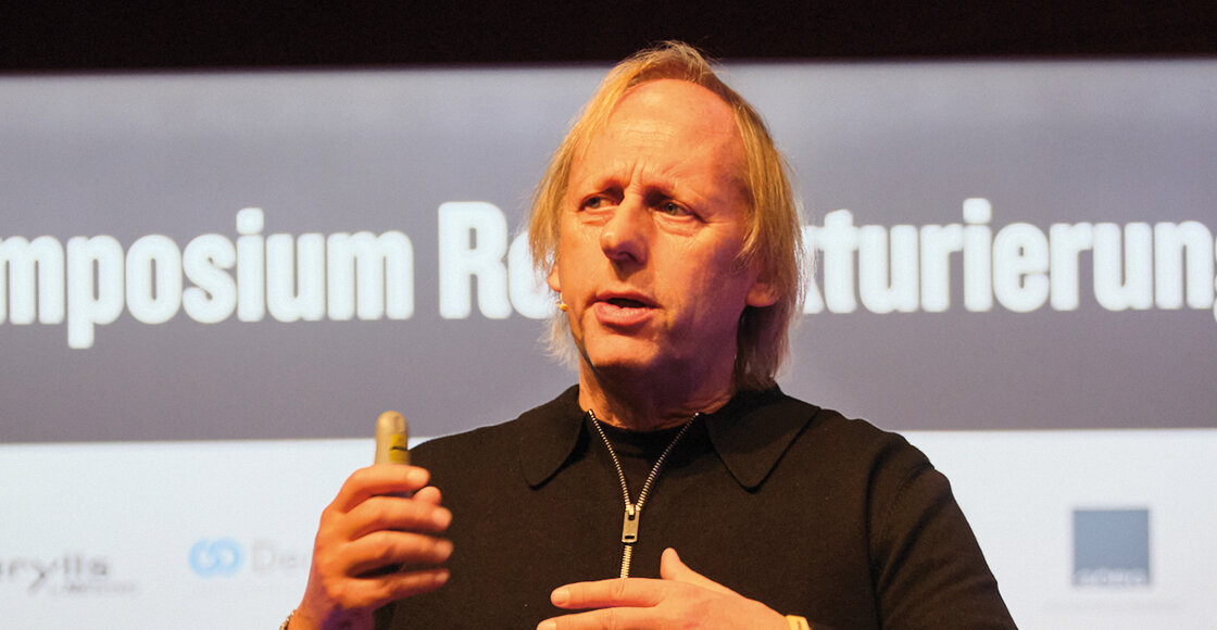 2024_Exler | © FH Kufstein Tirol Ein Mann spricht auf einer Bühne und hält ein Präsentationsgerät in der Hand, im Hintergrund sind der Schriftzug „Symposium Restrukturierung“ und einige Sponsorenlogos zu sehen. | © FH Kufstein Tirol