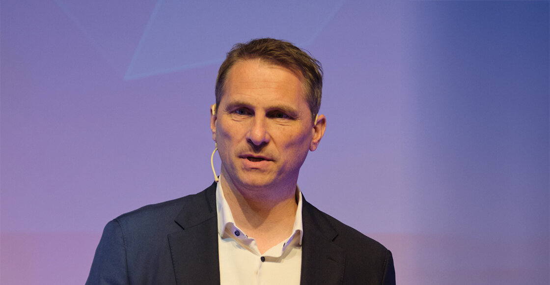 2024_Danninger_Oliver | © FH Kufstein Tirol Ein Mann in Anzug und Hemd spricht auf einer Bühne und trägt ein Mikrofon am Ohr, vor einem dezenten, lilafarbenen Hintergrund | © FH Kufstein Tirol