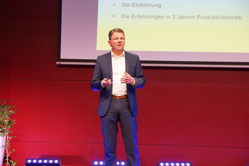 Ein Vortragender steht auf der Bühne. Hinter ihm befindet sich eine große Leinwand, auf der eine PowerPoint-Präsentation angezeigt wird. | © FH Kufstein Tirol