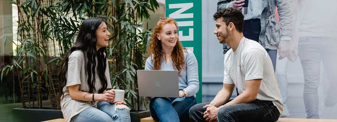 Drei Studierende der FH Kufstein Tirol sitzen zusammen in der Aula, unterhalten sich und arbeiten am Laptop.