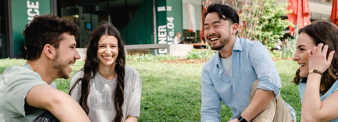 Studierende der FH Kufstein Tirol sitzen gemeinsam auf der Wiese am Campus und unterhalten sich in entspannter Atmosphäre.