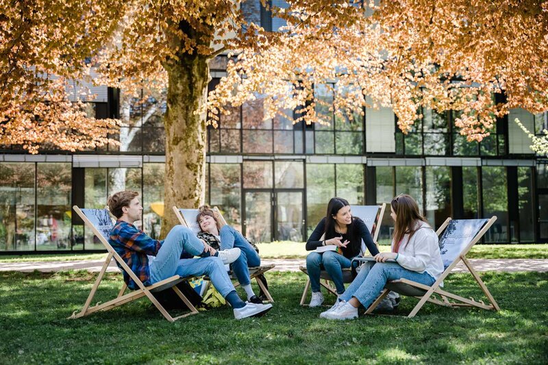 Vier Studierende der FH Kufstein Tirol sitzen im Park vor dem Campus in Liegestühlen und unterhalten sich bei sonnigem Wetter.