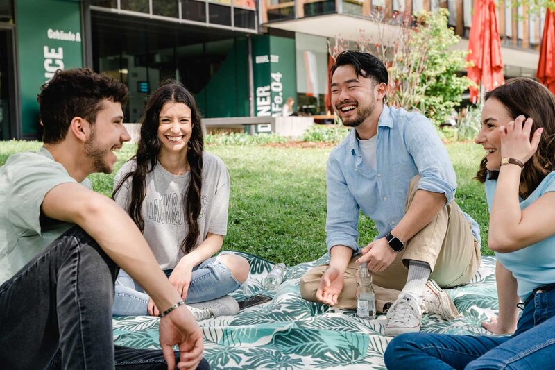 Studierende der FH Kufstein Tirol sitzen gemeinsam auf der Wiese am Campus und unterhalten sich in entspannter Atmosphäre.