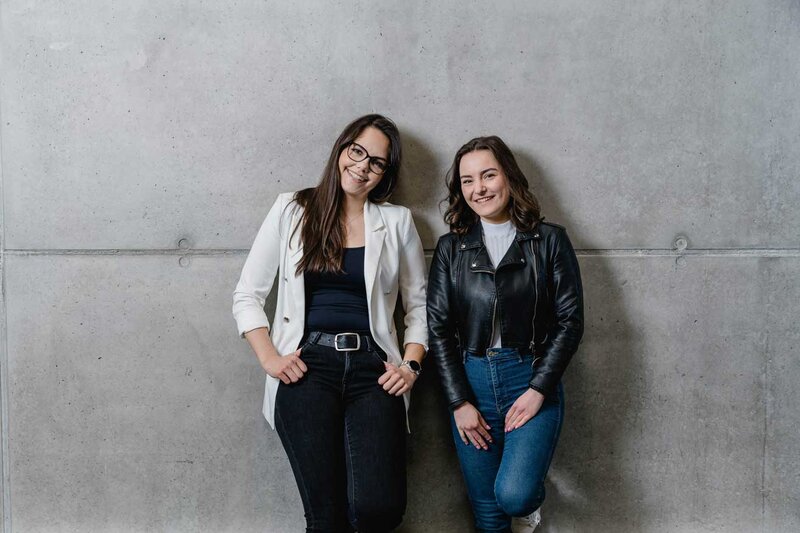 Zwei Studentinnen der FH Kufstein Tirol posieren lächelnd vor einer Betonwand am Campus.