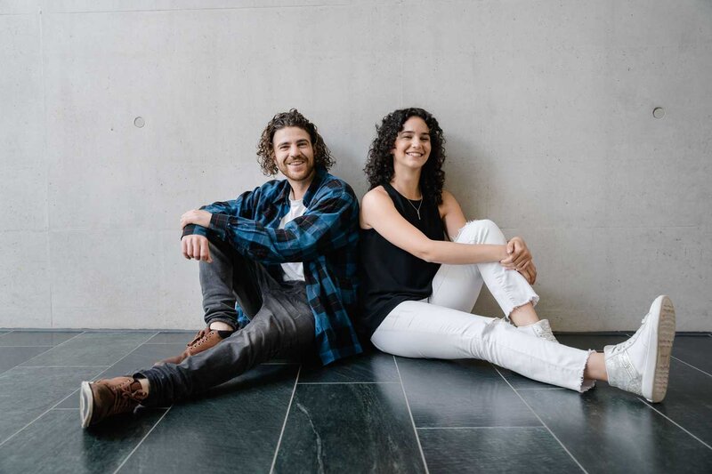 Zwei Studierende der FH Kufstein Tirol sitzen entspannt am Boden vor einer Betonwand und lächeln in die Kamera.