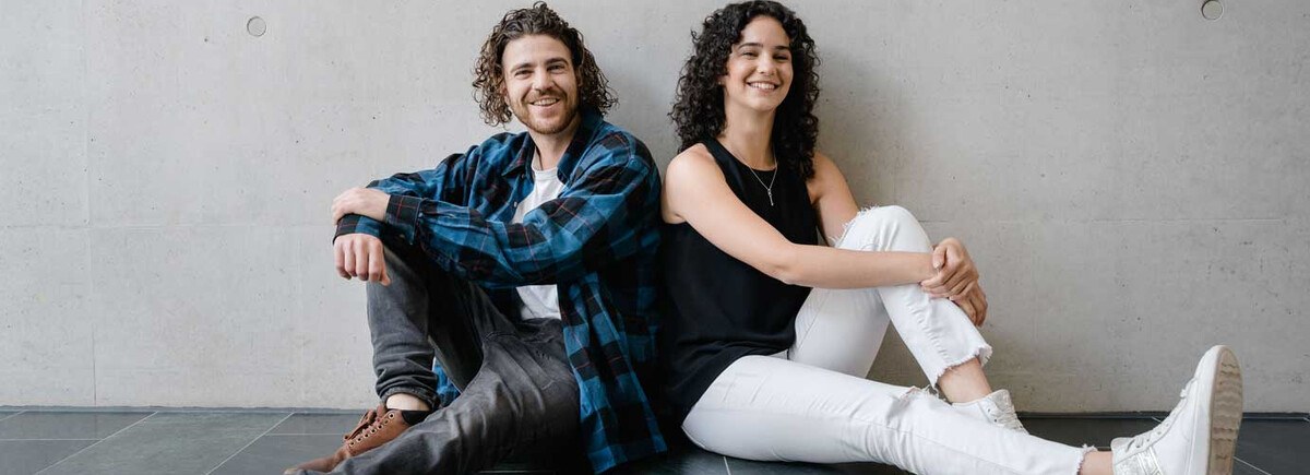 Zwei Studierende der FH Kufstein Tirol sitzen entspannt am Boden vor einer Betonwand und lächeln in die Kamera.