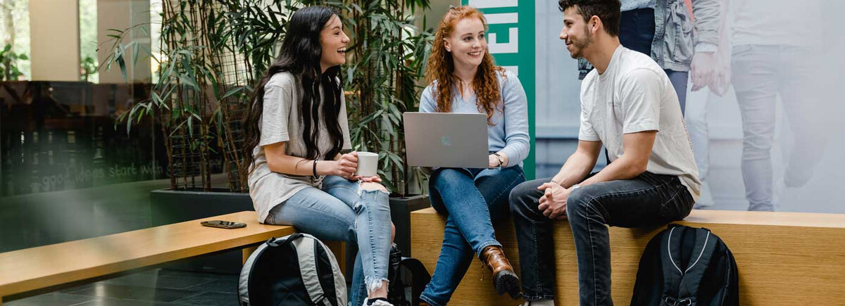 Drei Studierende der FH Kufstein Tirol sitzen zusammen in der Aula, unterhalten sich und arbeiten am Laptop.