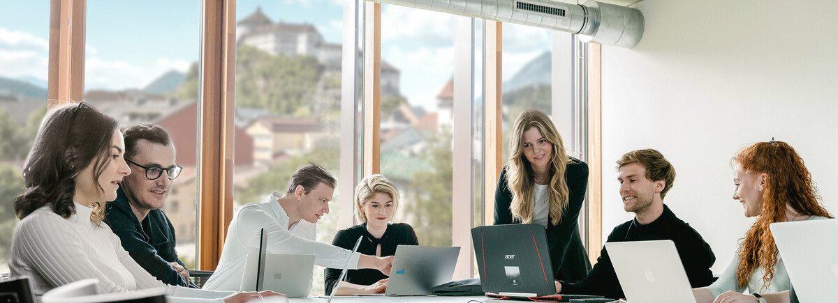 Studierende an der FH Kufstein Tirol sitzen an einem Tisch mit Laptops und diskutieren in einem hellen Raum mit Blick auf die Festung Kufstein.