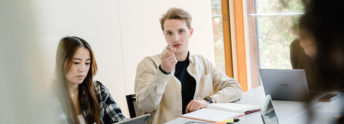 Language_Center | © FH Kufstein Tirol Drei Personen sitzen an einem Tisch, und ein Mann dieser Gruppe zeigt mit seinem Stift in die Richtung der Kamera, wo sich ein Whiteboard befindet. | © FH Kufstein Tirol