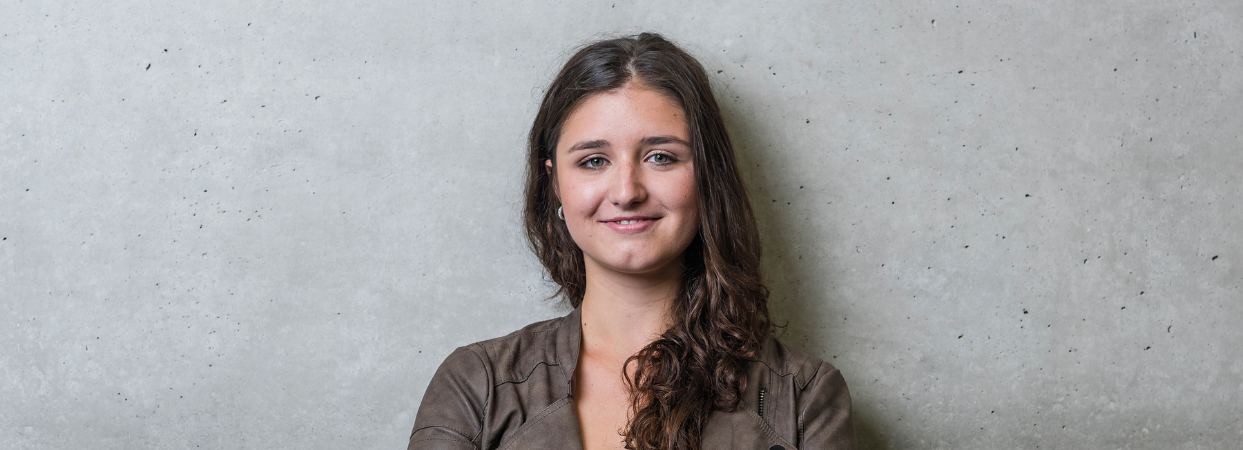 BA_IBS_VZ | © FH Kufstein Tirol Eine Frau steht mit dem Rücken zu einer grauen Wand und blickt lächelnd in die Kamera. | © FH Kufstein Tirol