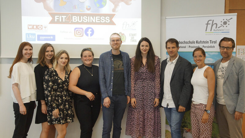 Das Jurorenteam des Wettbewerbs Fit4Business steht vor einer Leinwand auf dem der Schriftzug FH Kufstein Tirol zu lesen ist; an der zweiten Wand die Einblendung einer Präsentation zum Wettbewerb Fit4Busines | © FH Kufstein Tirol