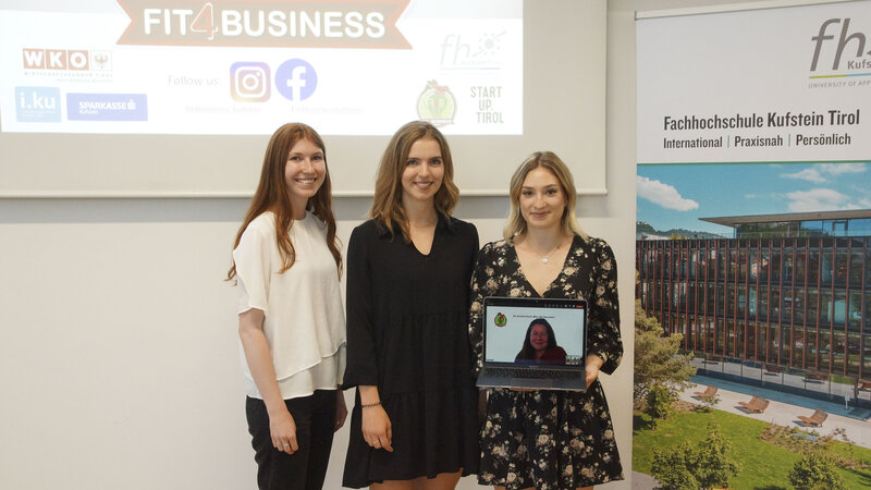Drei junge Frauen stehen von einer Wand auf der eine Präsentation gezeigt wird. Zu lesen ist der Schriftzug Fit4Business; auf der zweiten Wand ist ein Plakat der FH Kufstein Tirol aufgehängt; die jungen Frauen halten einen Laptop, darin sieht man den Screen einer Video-Call-Schaltung  | © FH Kufstein Tirol