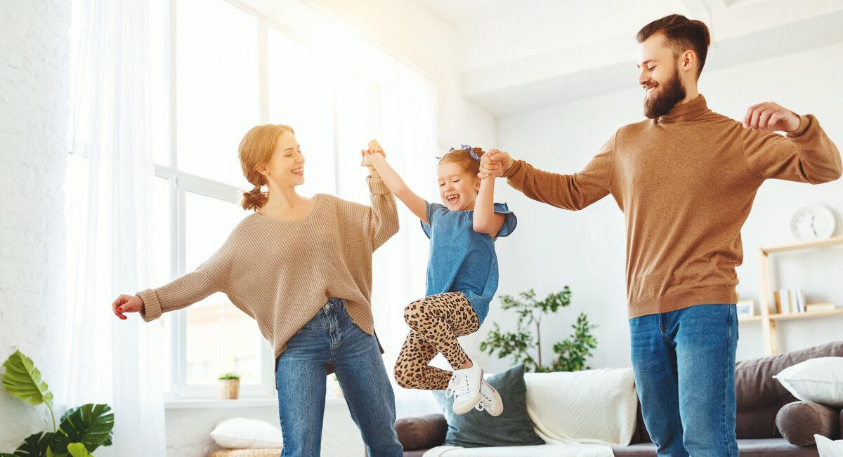 Eltern-Kind-froehlich-Zimmer | © AdobeStock.JenkoAtaman röhliche Eltern schwingen ihr Kind zwischen sich, während sie in einem sonnendurchfluteten Wohnzimmer stehen. Alle tragen lässige Freizeitkleidung und genießen den Moment gemeinsam. | © AdobeStock.JenkoAtaman