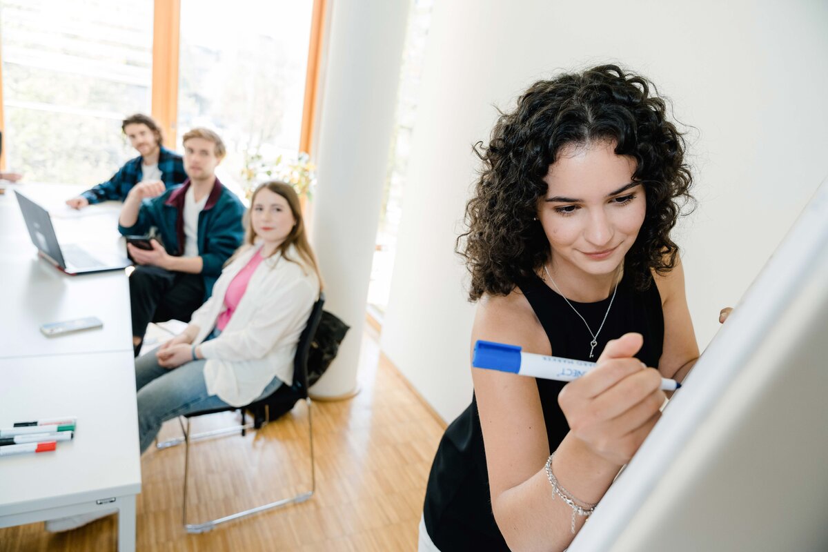 Gruppenprojekt_Fotoshooting_2023 | © FH Kufstein Tirol Junge Frau schreibt mit einem blauen Marker auf ein Whiteboard, während drei Studierende ihr von einem Tisch aus zuschauen. | © FH Kufstein Tirol