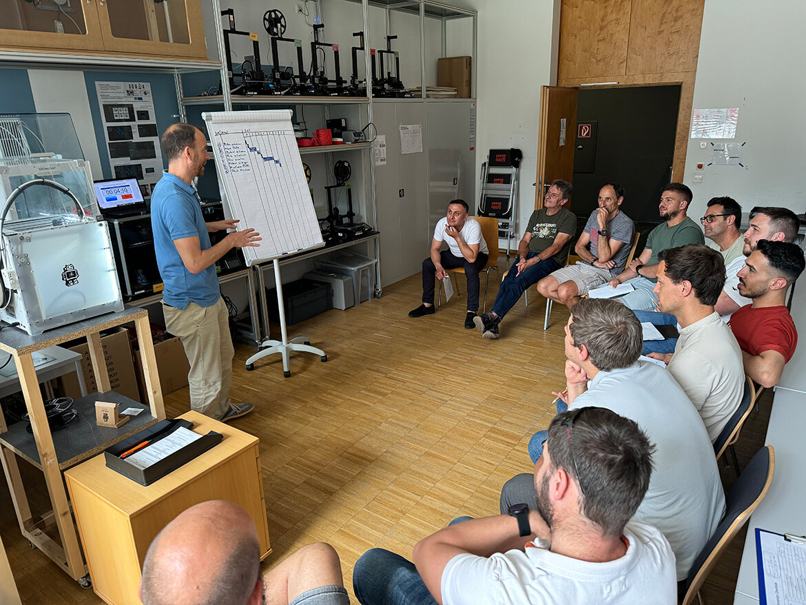 SMED-Training Lean Lab | © FH Kufstein Tirol Männer sitzen auf Stühlen in einem Kreis und schauen zu einem Mann im blauen Polo-Hemd, der vor einem Flipchart steht. | © FH Kufstein Tirol