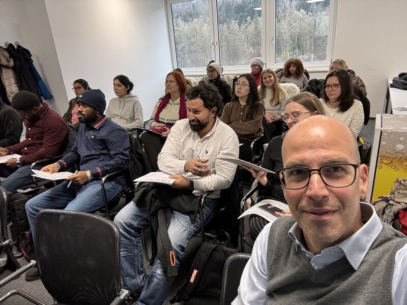 Ein Mann mit Glatze sitz vor einer Gruppe von Menschen auf einem Stuhl und macht ein Selfie. | © FH Kufstein Tirol/FERCAM