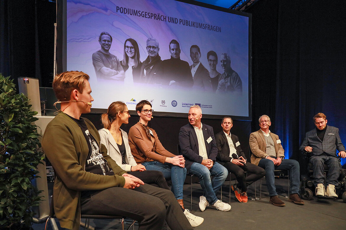 Kongress Resilienter Tourismus Speaker | © Chiemgau GmbH – Tourismus Personen sitzen auf einem Podium auf einer Bühne vor einem Bildschirm | © Chiemgau GmbH – Tourismus